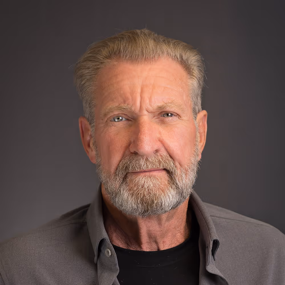Mature man with grey hair and beard wearing a dark shirt against a grey background.