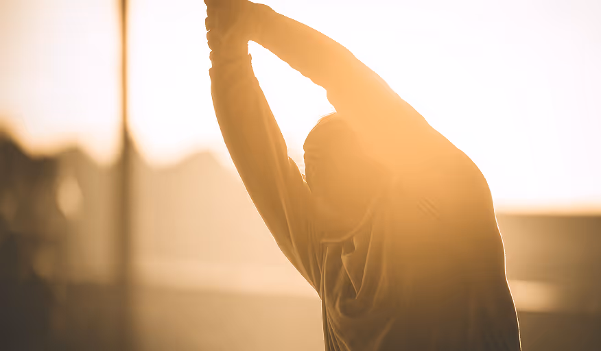 Person stretching arms upward in warm sunlight with blurred background.